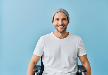 Cheerful disabled man seated in a wheelchair, wearing a white t shirt and gray beanie, smiling confidently at the camera against a bright blue studio background. concepts, accessibility, recovery, positive attitude, disability.の素材