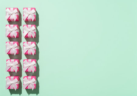 Ten bright pink miniature gift boxes with white ribbons arranged in two columns on a light mint green surface. minimalist flat lay with ample copy space for text or design.の素材