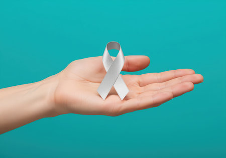 Silver awareness ribbon, symbolizing support and solidarity, resting neatly on the open palm of a hand. isolated on a vibrant teal studio background. concept of health, medicine, and disease awareness.の素材
