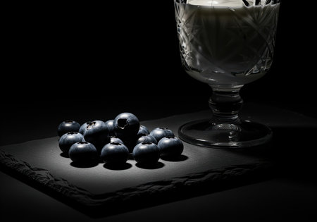 Dark blueberries piled on a rough slate serving board, dramatically lit in a low key studio setting, positioned next to an ornate crystal glass filled with white dairy product.の素材