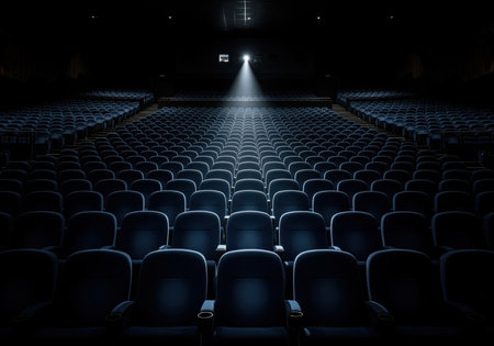 Empty rows of dark blue seats in a large, dark cinema auditorium. a single, powerful beam of light cuts through the darkness from the projector above, creating a dramatic atmosphere.の素材