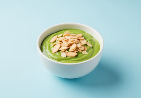 Vibrant green creamy dessert, possibly matcha yogurt or pudding, served in a white bowl and garnished with sliced almonds on a light blue surface. healthy food concept.の素材