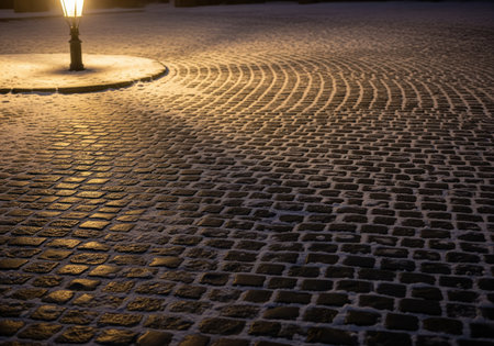 Close up view of a historic cobblestone path lightly covered in snow, illuminated by the warm, golden glow of a vintage street lamp at night.の素材