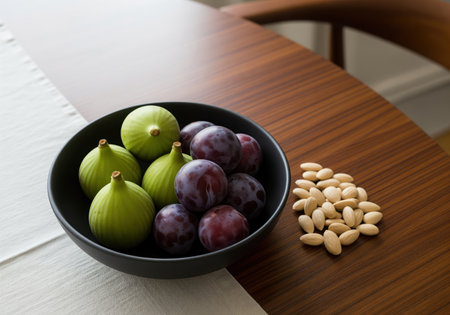 Green figs and purple plums arranged in a matte black bowl, accompanied by a pile of peeled almonds, resting on a polished wooden table.の素材