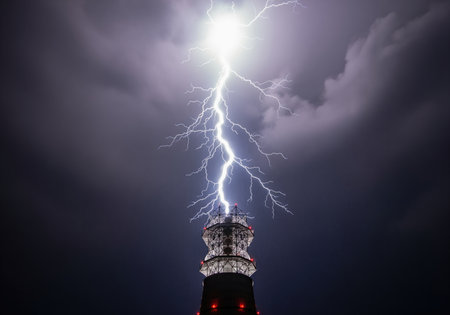 Powerful electrical discharge of white lightning striking the top of a tall telecommunication tower against a dark, stormy night sky. dramatic weather phenomenon.の素材