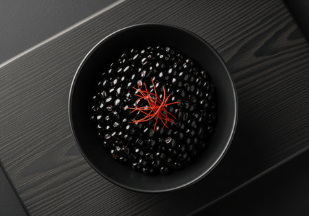 Black caviar, a luxurious delicacy, presented in a sleek black bowl on a dark wooden surface. garnished with delicate red threads. gourmet food concept.の素材