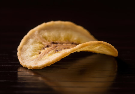 Golden crispy banana chip, a healthy dried fruit snack, presented in a detailed macro studio shot on a dark reflective surface, emphasizing texture.の素材