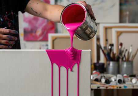 Thick, vibrant magenta paint is poured from a metal can, dripping down the side of a white canvas in an artist studio. creative process, fluid motion, and abstract art technique.の素材