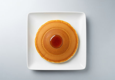 Single, thick, perfectly circular golden pancake centered on a square white plate, featuring a small, glossy drop of maple syrup in the middle. clean studio shot, top view.の素材