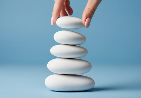 Woman hand carefully places the final smooth white stone atop a perfectly balanced stack of five rocks. studio shot emphasizing stability, zen, and tranquility against a clean blue background.の素材