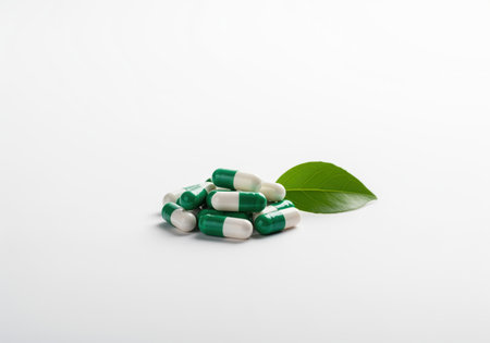 Green and white medical capsules piled neatly next to a fresh green leaf on a clean white background. concept of natural medicine, herbal supplements, and pharmaceutical wellness.の素材