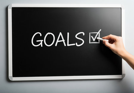 Hand drawing a white chalk checkmark inside a box next to the capitalized word GOALS written on a black chalkboard. represents achievement, planning, motivation, and business success.の素材