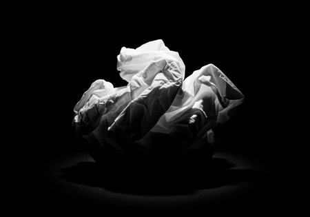 Crumpled white tissue illuminated by a harsh spotlight, casting dramatic shadows against a completely black background. abstract concept of stress, waste, or forgotten ideas.の素材