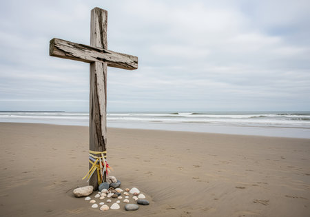 Weathered wooden cross memorial stands on a sandy beach with ocean waves under a cloudy sky. rocks and seashells at its base symbolize remembrance and loss.の素材
