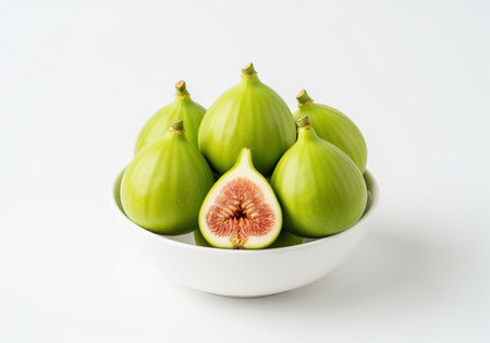 Vibrant green figs, one perfectly halved to reveal its red interior, are neatly arranged in a simple white ceramic bowl. fresh, healthy fruit on a clean, bright background.の素材