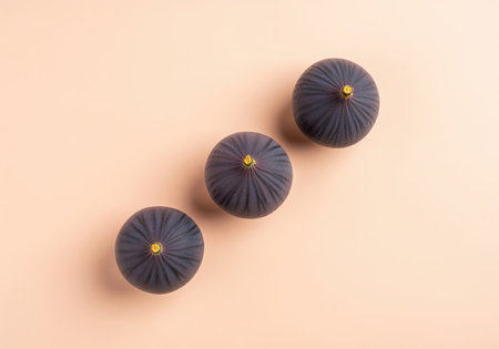 Whole dark purple figs with small yellow stems, neatly arranged in a diagonal row on a smooth, light peach background. perfect for healthy eating, fresh fruit, and natural food concepts.の素材