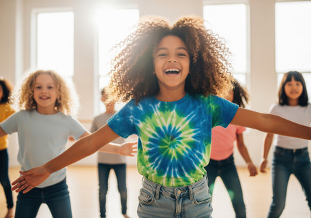 A joyful curly haired girl in a vibrant tiedye shirt smiles broadly while dancing with outstretched arms. she is surrounded by other diverse children in a bright studio, all engaged in energetic movement and fun.の素材