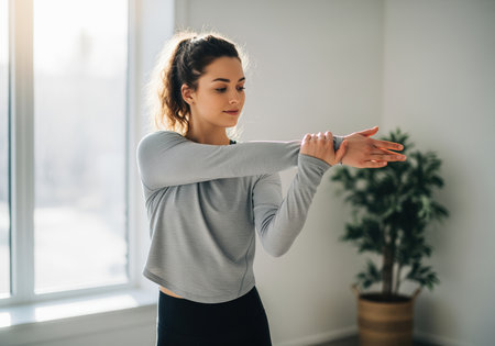 A young woman with a ponytail in athletic wear performs an arm stretch, focusing on her fitness routine. ideal for health and wellness content.の素材
