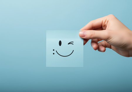 A person hand presents a light blue sticky note featuring a hand drawn winking smiley face, conveying happiness and positive emotion.の素材