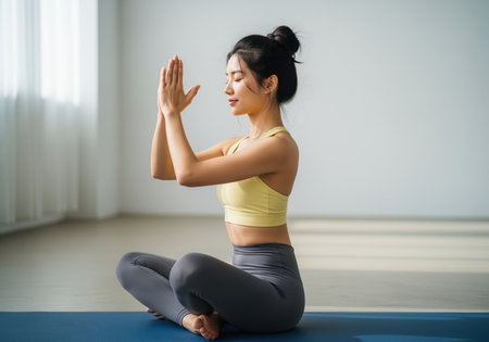 A serene young asian woman sits cross legged on a blue yoga mat, eyes closed, hands in prayer position, practicing meditation or mindfulness. this image conveys peace and wellness.の素材