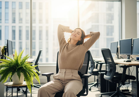 A content young woman takes a mindful break, stretching her arms behind her head while seated in her office chair, enjoying the sunlight. this image conveys relaxation and well being in a modern work environment.の素材