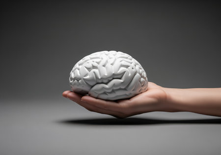 A human hand gently cradles a detailed white anatomical model of a brain, symbolizing intelligence, health, and thought, set against a simple gray background.の素材