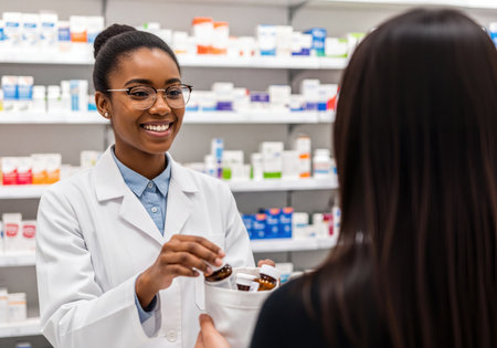 Happy African American pharmacist in a white coat and glasses hands bottles of medication to a customer at a pharmacy counter. Shelves with various medicines line the background.の素材