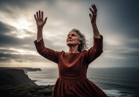 Elderly woman standing on a rugged cliff with arms raised towards a dramatic, cloudy sky, overlooking the vast ocean. Evokes feelings of hope, prayer, and freedom.の素材