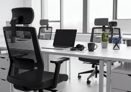 Modern office interior showcasing a clean workspace with an empty desk, laptop, ergonomic office chair, headphones, coffee mug, and cactus. Bright natural light fills the contemporary corporate environment.の素材