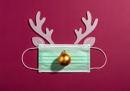 A surgical face mask is creatively transformed into a Christmas reindeer with white antlers, ears, and a shiny golden bauble for a nose, set against a deep red background. Represents Holiday season during a pandemic.の素材