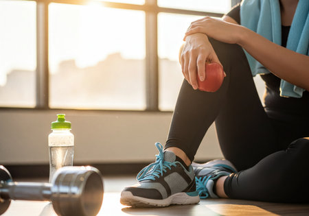 Woman paused during a workout, holding a red apple, sitting near a water bottle and dumbbell. Emphasizes healthy lifestyle, sport, and nutrition indoors.の素材