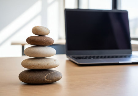 Stacked zen stones on a light wooden office desk next to a modern laptop. Symbolizing work life balance, harmony, stability, and peaceful productivity in a business environment.の素材