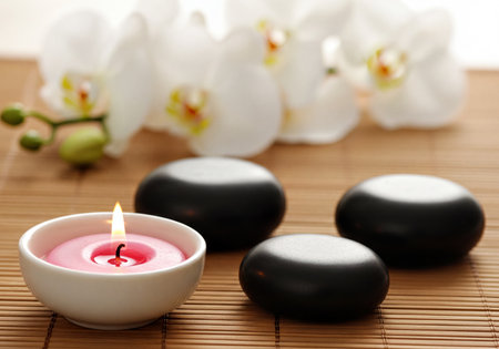 Burning pink candle in a white bowl by smooth black spa stones and delicate white orchid flowers arranged on a natural bamboo mat, perfect for relaxation and wellness concepts.の素材
