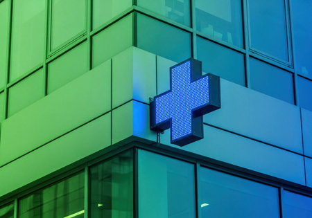 Bright blue LED cross sign mounted on the exterior corner of a modern architectural glass building, often indicating a pharmacy, hospital, or medical service location. Illuminated symbol against contemporary facade.の素材