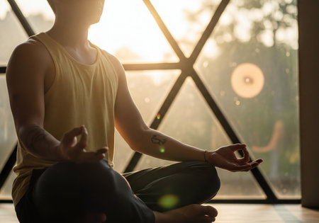 Man sits cross-legged in a peaceful lotus pose, hands in a mudra gesture, meditating by a sun-drenched window with geometric frames. Golden light floods the scene, evoking tranquility, mindfulness, and a healthy lifestyle.の素材