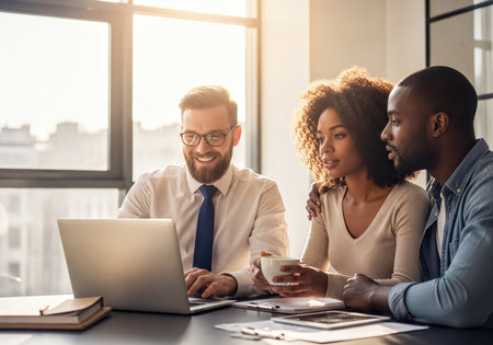 Diverse business team collaborating and discussing work around a laptop in a luminous modern office with city views, bathed in warm sunlight.の素材