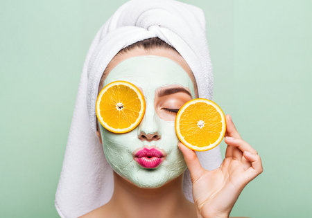 Young woman wearing a purifying green clay facial mask, holding fresh orange slices over her eyes with a towel wrap, promoting natural beauty and spa skincare.の素材