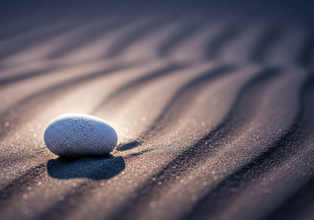 Smooth grey stone on fine, textured sand showing distinct wave patterns. Soft light illuminates the sand, creating sparkling highlights and subtle shadows with a bokeh effect. Focus on tranquility and minimalism.の素材