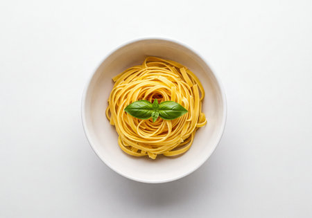 Raw tagliatelle pasta coiled neatly into a nest shape, placed in a minimalist white bowl and garnished with a single vibrant green basil leaf. Set against a clean, bright white background from an overhead perspective, emphasizing simple, fresh ingredients, Italian cuisine, and preparation.の素材