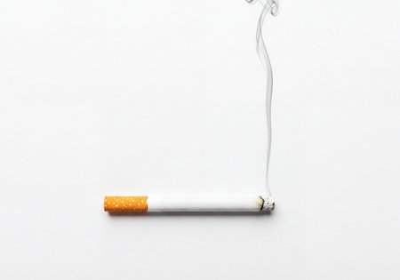 A single lit cigarette burning horizontally on a pristine white surface, a thin plume of smoke gracefully ascending from its glowing ember and ash.の素材