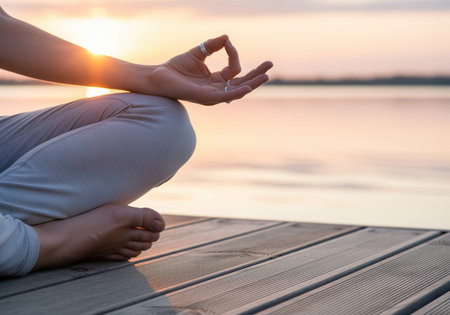 An individual sits in a meditative lotus position on a wooden dock near water at sunset, showing the hand in the gyan mudra gesture, symbolizing focus, peace, and spiritual wellness.の素材