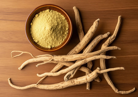 Dried indian ginseng ashwagandha roots are piled next to a small ceramic bowl filled with fine herbal powder, resting on a rich brown wooden surface, representing traditional ayurvedic medicine and natural health supplements.の素材