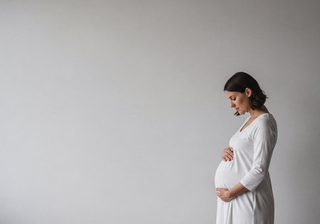 Pregnant woman in white dress standing in profile, gently cradling her belly with both hands, looking down thoughtfully against a simple light background. Concept of motherhood, anticipation, new life, and tenderness.の素材