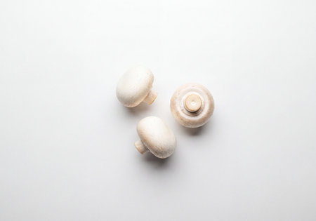 Three raw white button mushrooms, also known as champignon mushrooms, arranged on a pristine white surface. This clear top-down view highlights their fresh texture, ideal for culinary projects, healthy eating, and ingredient photography.の素材