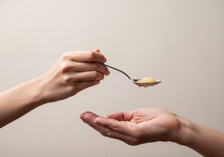 A child's hand holds a spoon with pureed food, extending it towards an adult's open palm, illustrating feeding, giving, and receiving in a moment of care and nourishment.の素材
