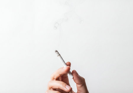 Close up of a human hand holding a lit cigarette with a burning tip and visible smoke ascending into a clean white background. Represents concepts of smoking, addiction, and health risks.の素材