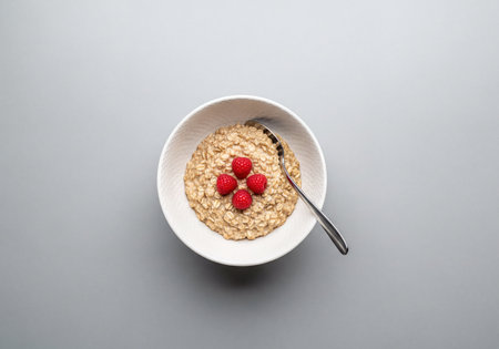 Bowl of warm oatmeal porridge topped with fresh red raspberries, presented with a silver spoon on a clean gray surface. A wholesome and nutritious breakfast.の素材