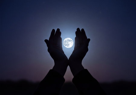 Two silhouetted hands appear to hold a luminous full moon against a clear, gradient dark blue and purple night sky. The brilliant sphere of the moon glows brightly, centered between the fingertips, symbolizing wonder and connection.の素材