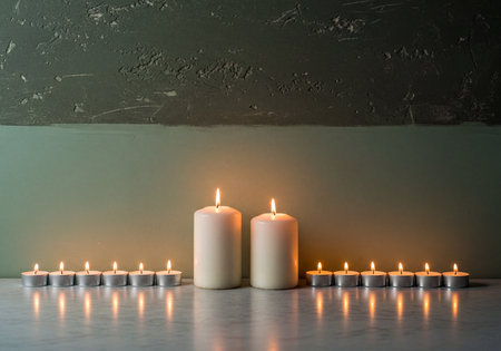Two large pillar candles and a row of small tealight candles burn brightly on a reflective surface, symmetrically arranged against a textured two toned wall, creating a warm and serene atmosphere.の素材