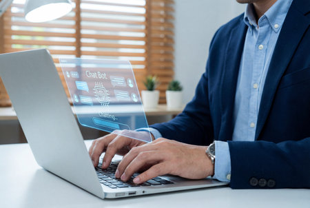 A professional man in a blue suit types on a laptop while a futuristic blue holographic chat bot interface floats above the screen displaying digital messages and artificial intelligence icons.の素材